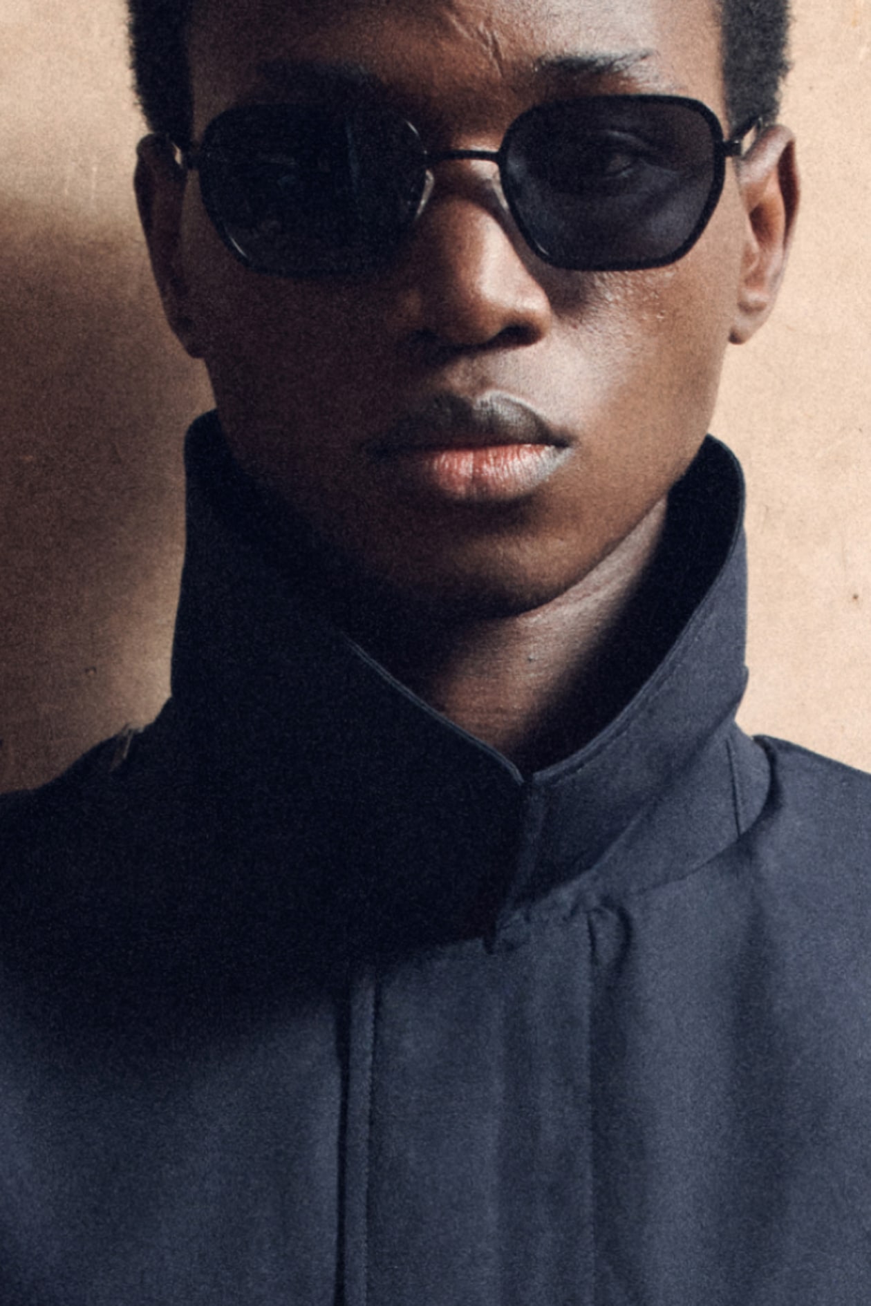 Person wearing dark sunglasses and a high-collared jacket, with a neutral expression, facing the camera against a plain background.