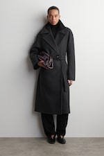 Placeholder for: Person wearing a long, dark coat with a belt, black pants, and holding a dark, glossy clutch against a plain white background.