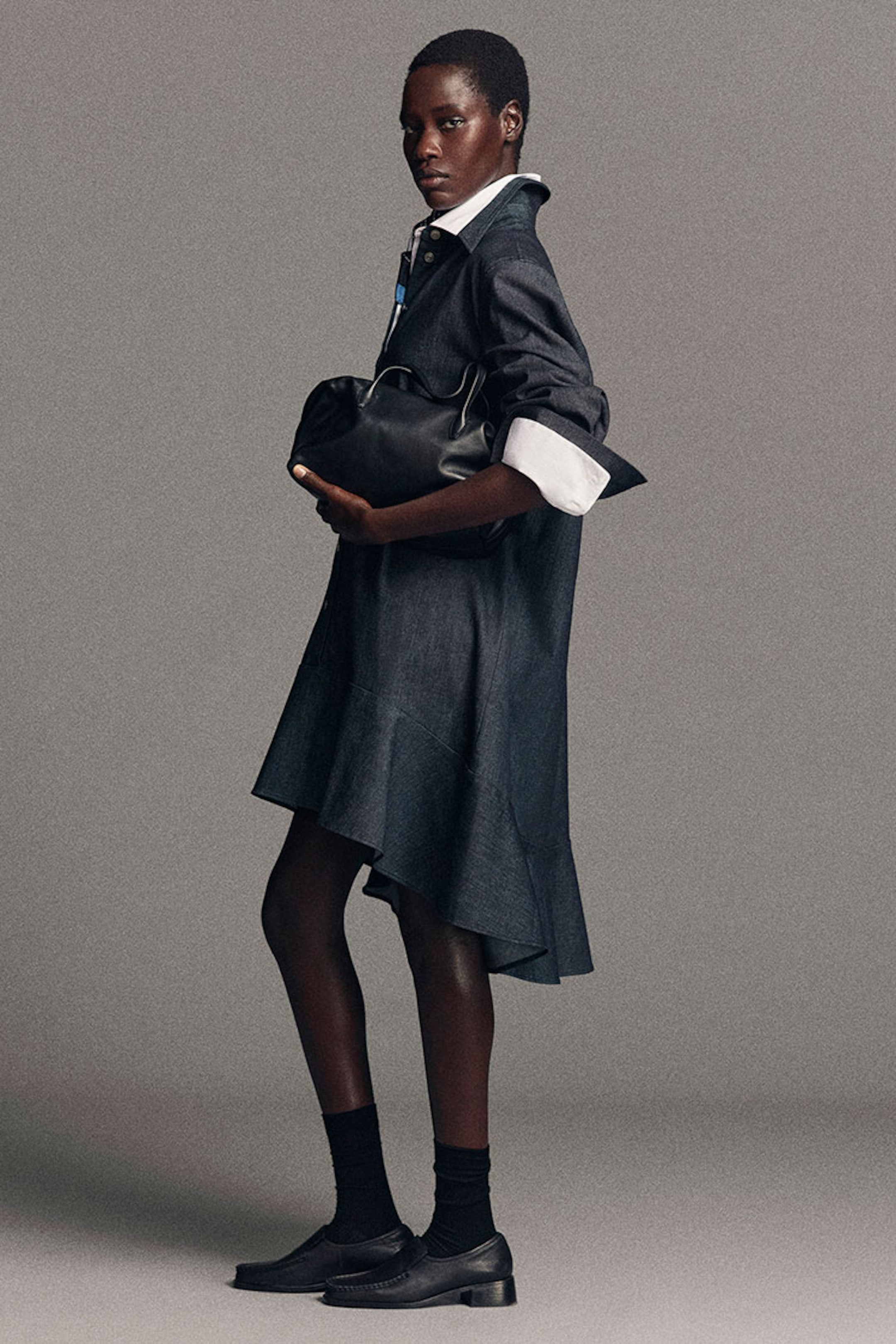 Person in a denim dress with white cuffs holds a black handbag, standing against a gray background, wearing black socks and shoes.