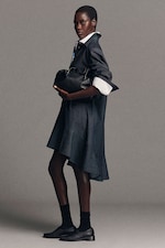 Placeholder for: Person in a denim dress with white cuffs holds a black handbag, standing against a gray background, wearing black socks and shoes.