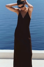 Placeholder for: Person in a sleeveless black dress poses by the sea, wearing a headband and sunglasses, with arms raised, under a clear blue sky.