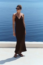Placeholder for: A person wearing a black dress and sunglasses stands near calm, blue water on a sunny day.