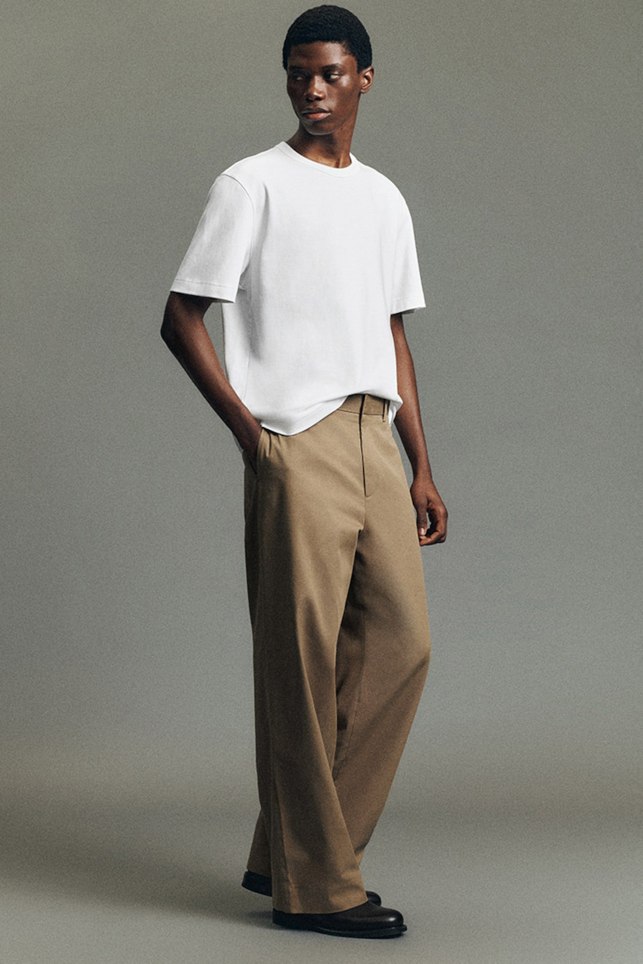 Person wearing a white T-shirt, beige trousers, and black shoes, with hands in pockets, standing against a neutral gray background.