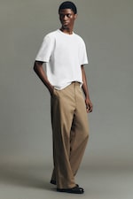 Placeholder for: Person wearing a white T-shirt, beige trousers, and black shoes, with hands in pockets, standing against a neutral gray background.