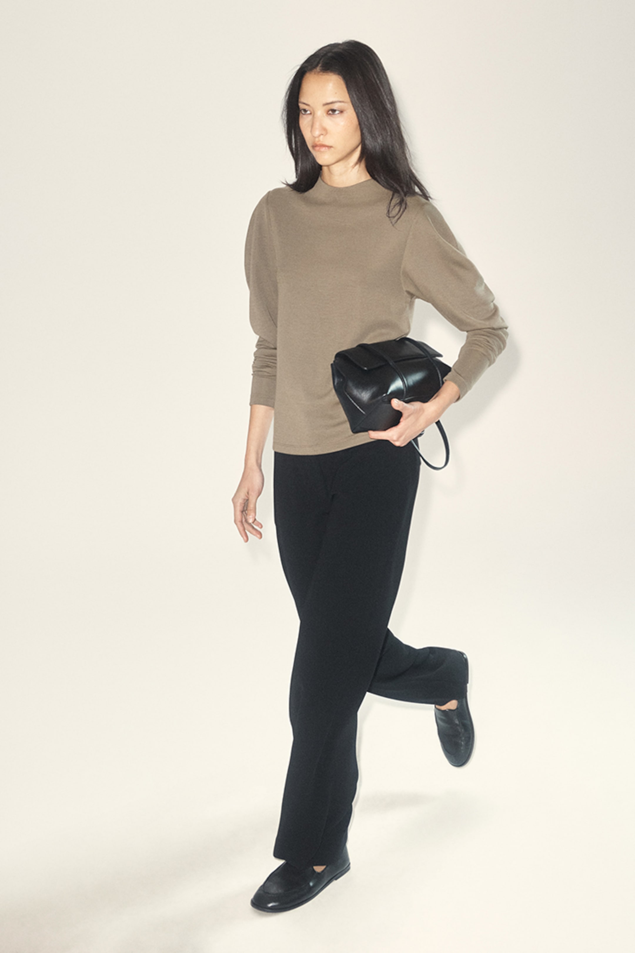 A woman in a tan sweater and black pants walks, holding a black bag. She has long dark hair and is wearing black shoes.