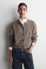 Placeholder for: Person wearing a brown button-up sweater over a white blouse and dark jeans, standing against a light background.