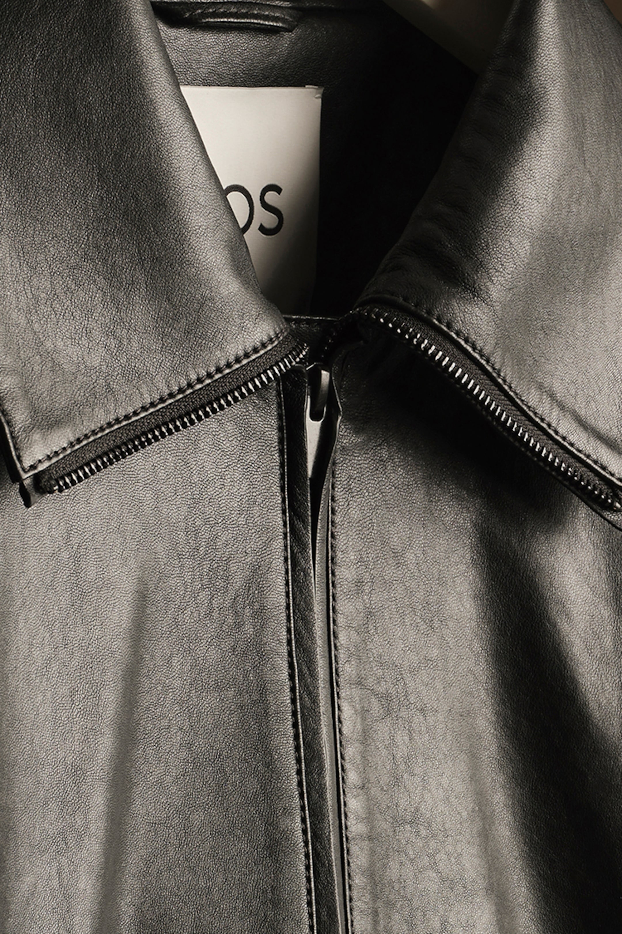 Close-up of a black leather jacket with a visible zipper and collar, hanging on a hanger. A label is partially visible inside.