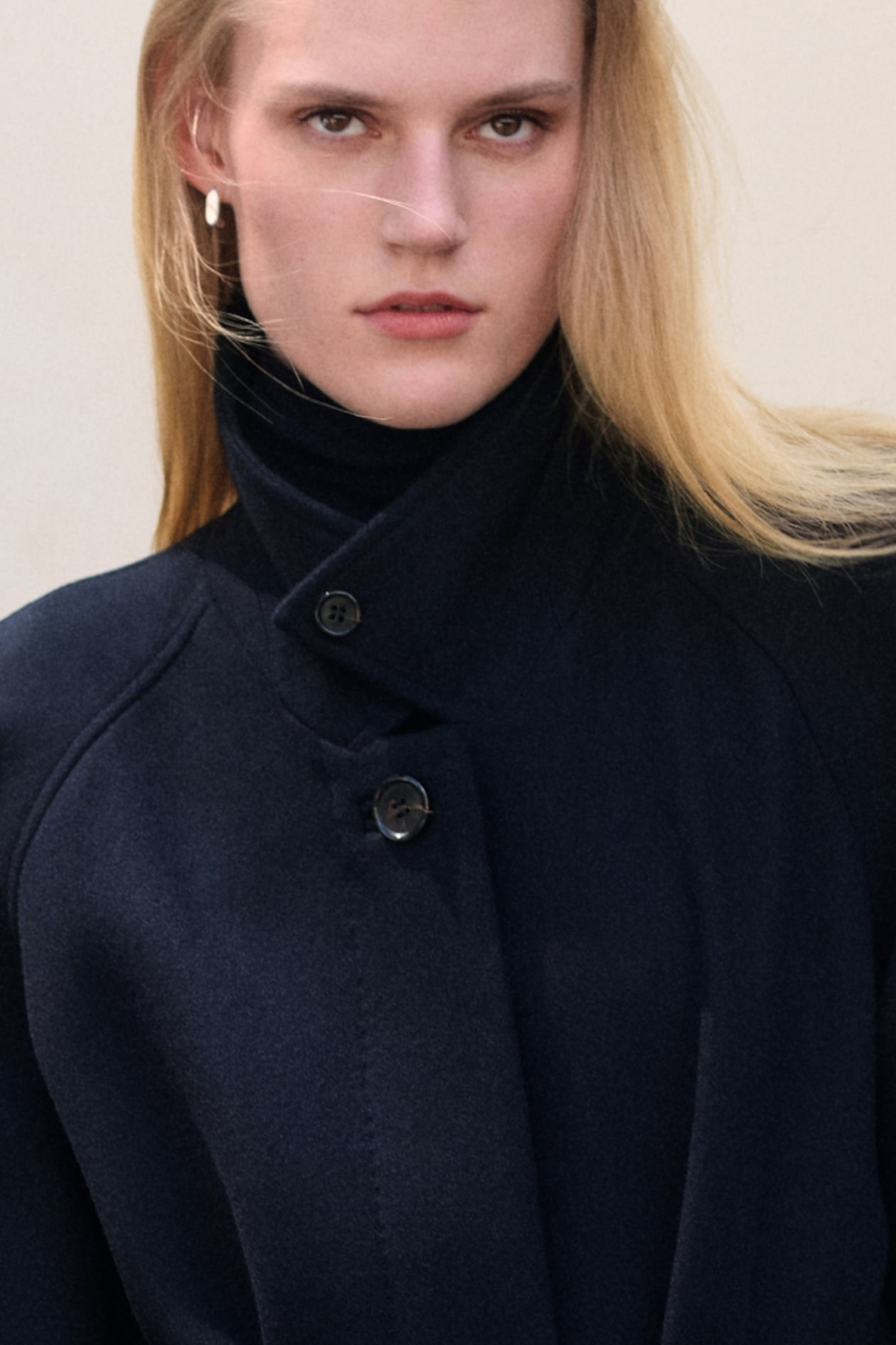 Person with long blonde hair wearing a dark high-collared coat, looking directly at the camera against a neutral background.