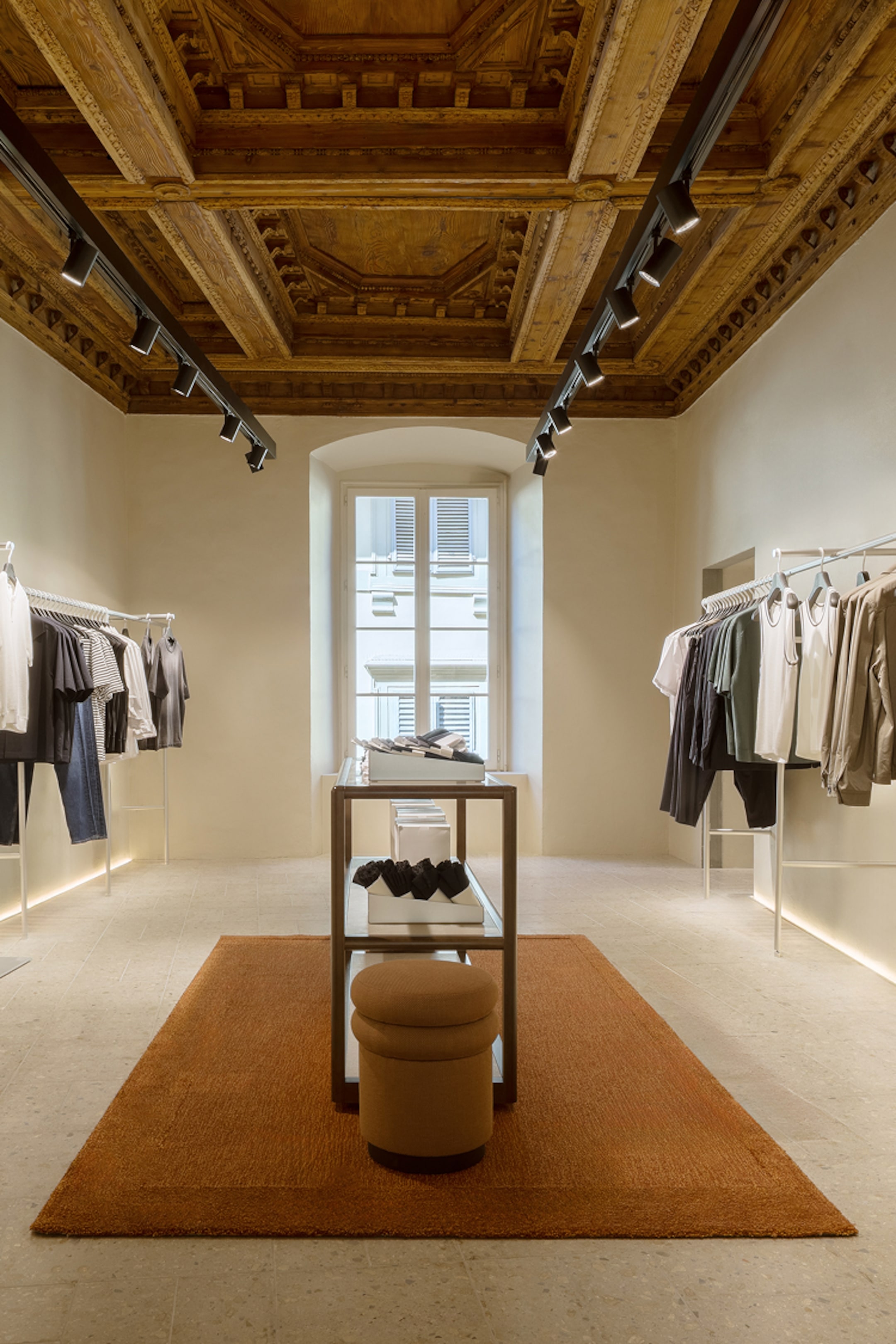 Clothing store with elegant wooden ceiling, racks of clothes, central display table, and a large window providing natural light.