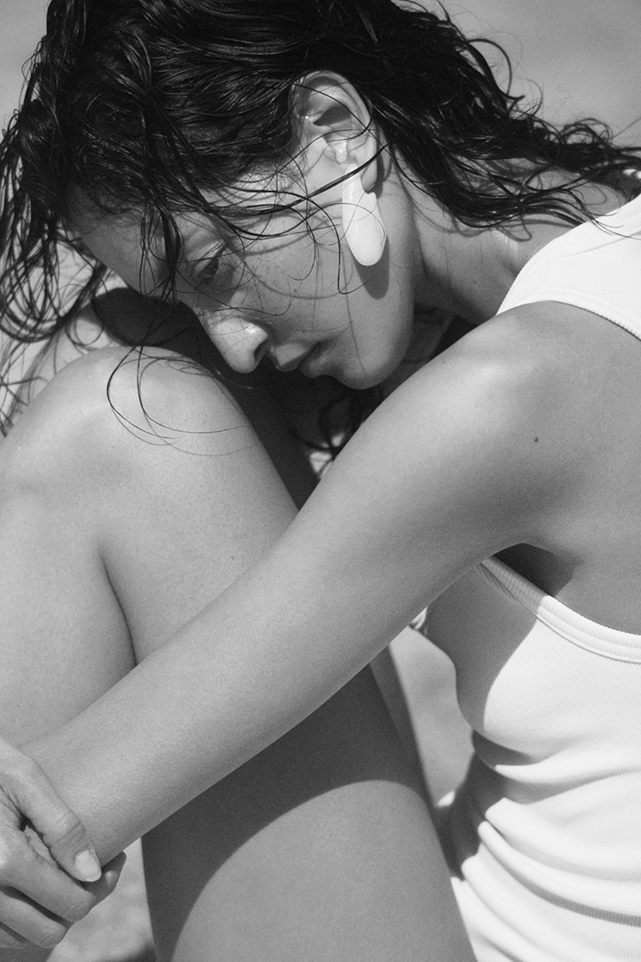 Black and white photo of a person with wet hair, wearing a tank top, sitting with knees drawn up, gazing downward.