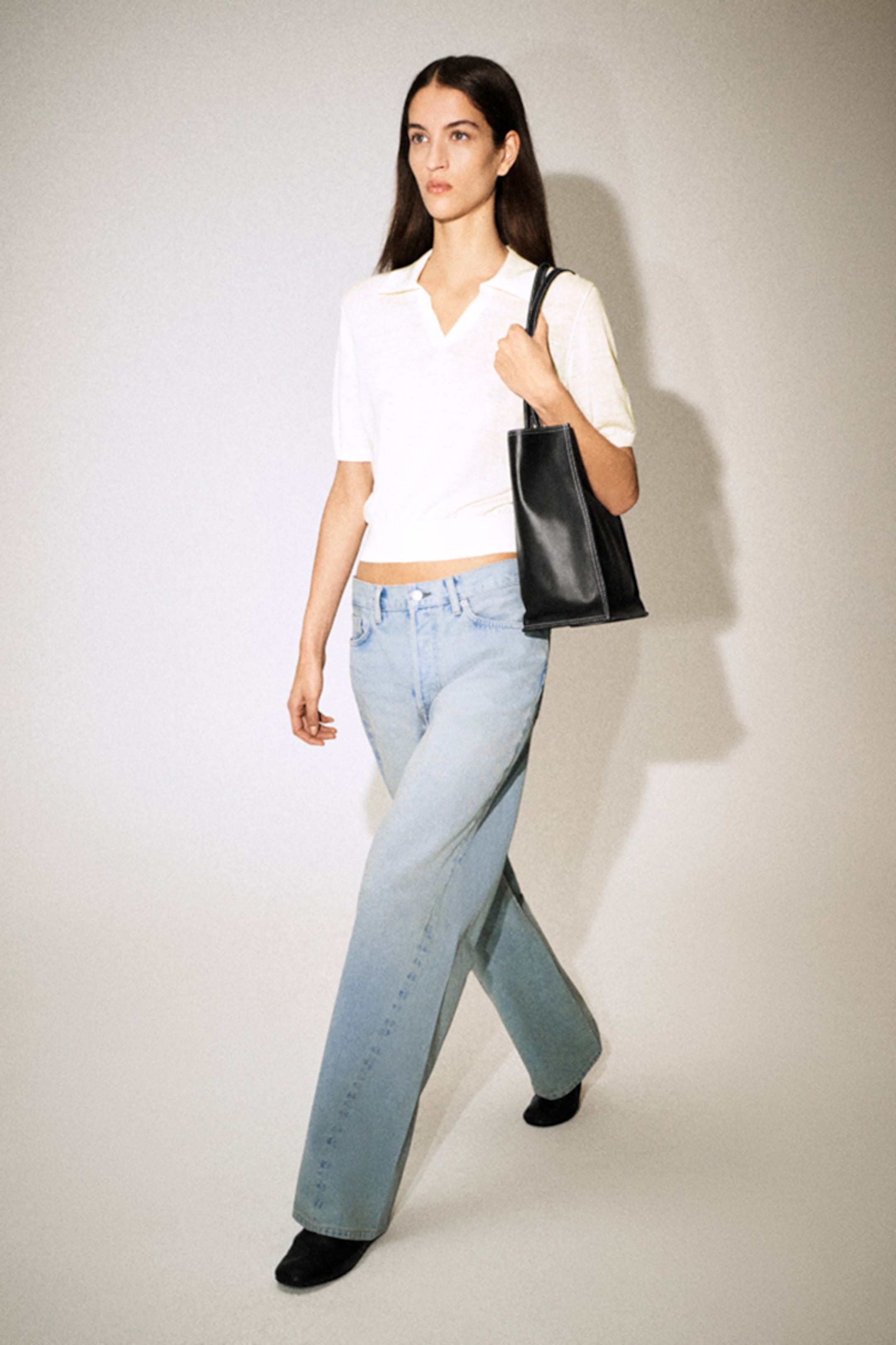 A woman with long hair walks confidently, wearing a white top, light blue jeans, and black boots. She carries a black handbag.