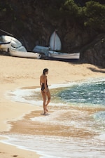 Placeholder for: A woman in a black swimsuit stands at the edge of the sea on a sandy beach, with boats and trees in the background.