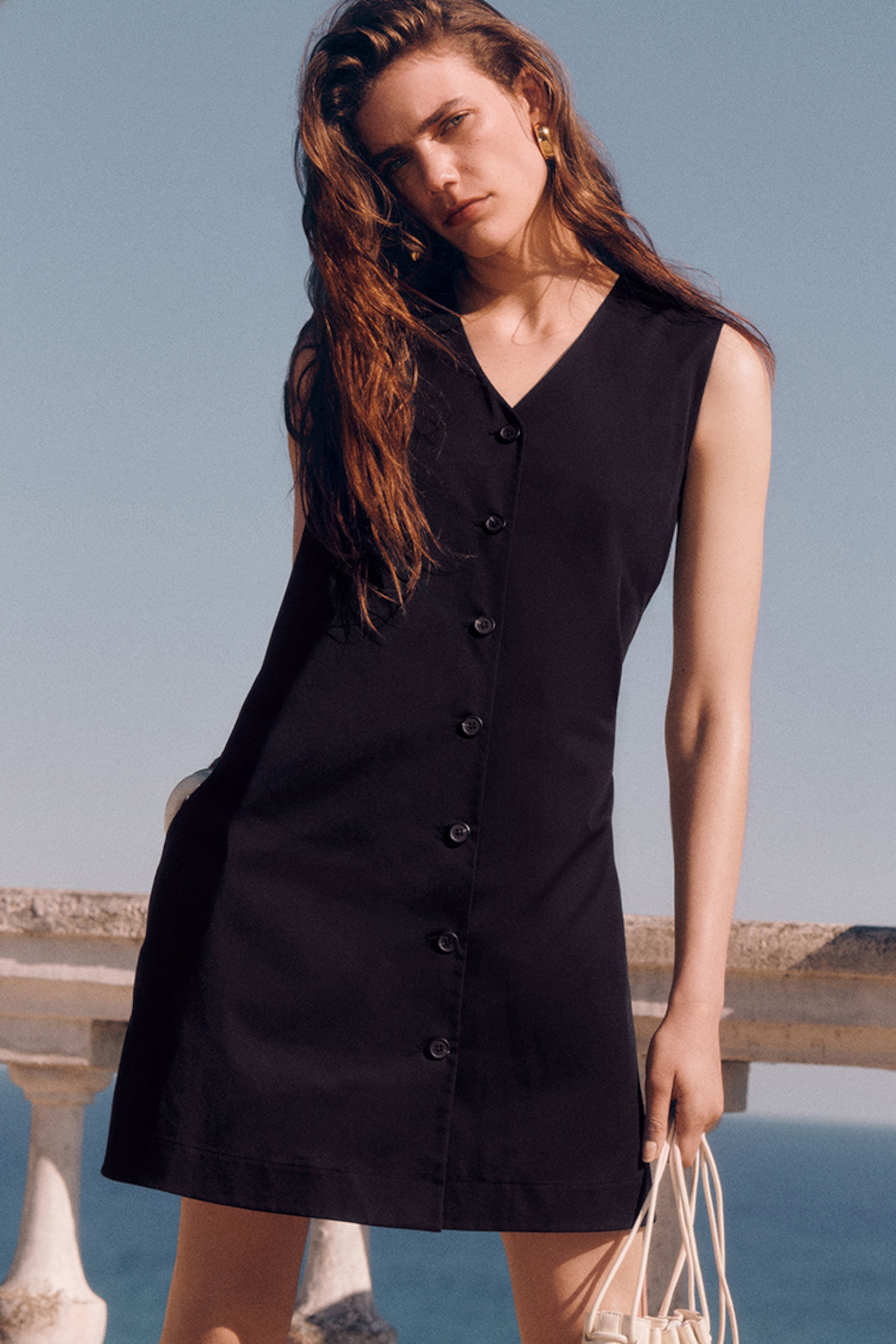 Woman in a sleeveless black dress poses outdoors, holding a light-colored bag. She stands against a clear blue sky and stone railing.