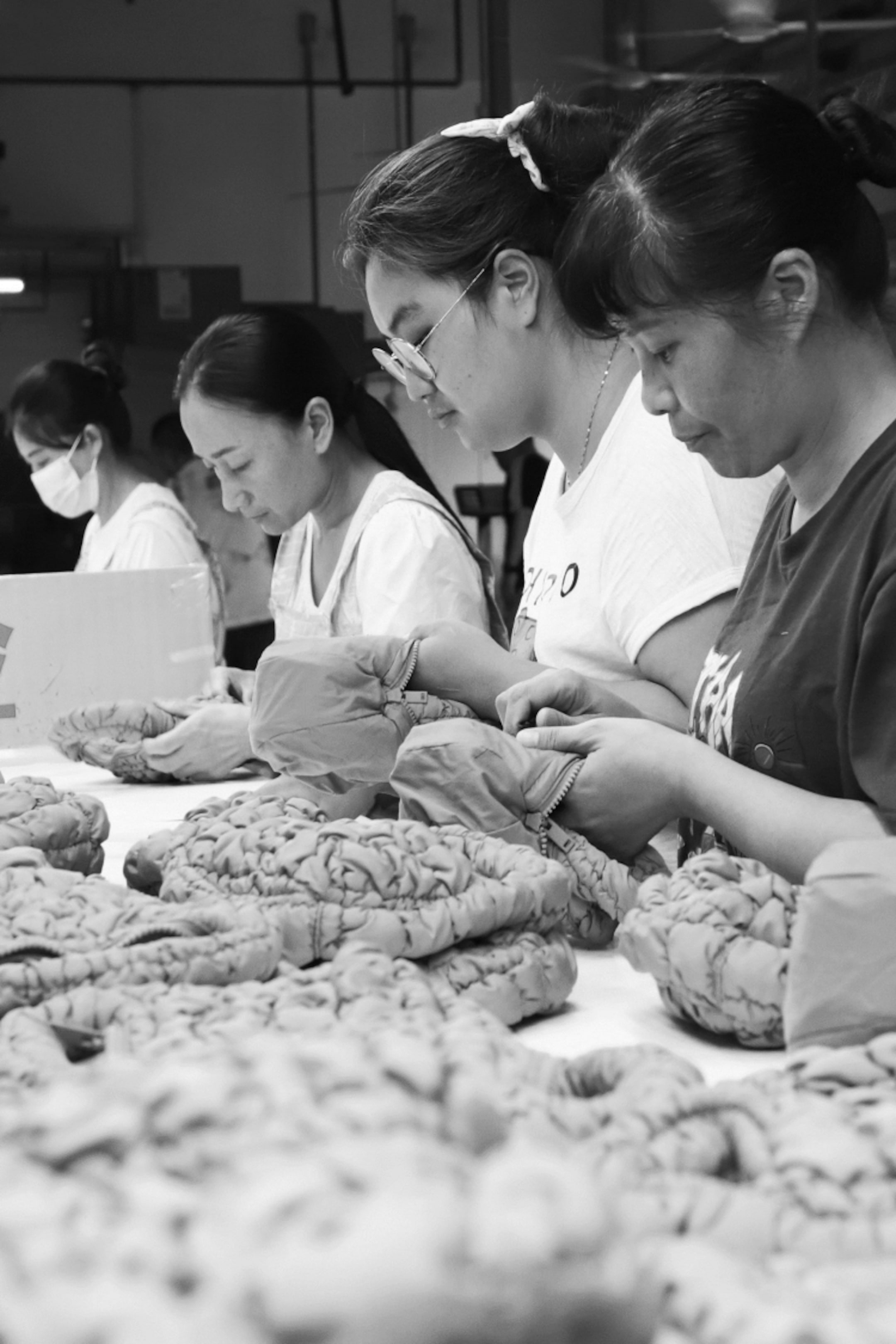 Four women focused on sewing fabric pieces at a table, surrounded by coiled textile materials in a workshop setting. Black and white photo.