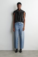 Placeholder for: Person wearing a dark top with a sweater tied around their shoulders, loose blue jeans, and black shoes, standing against a plain wall.