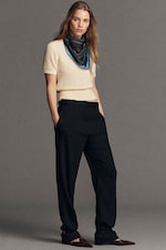 Placeholder for: Person in a cream sweater with a layered necklace, black wide-legged pants, and brown pointed shoes, standing against a gray background.