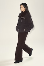 Placeholder for: Person walking in profile, wearing a dark brown jacket and pants, with hands in pockets, set against a plain, light background.