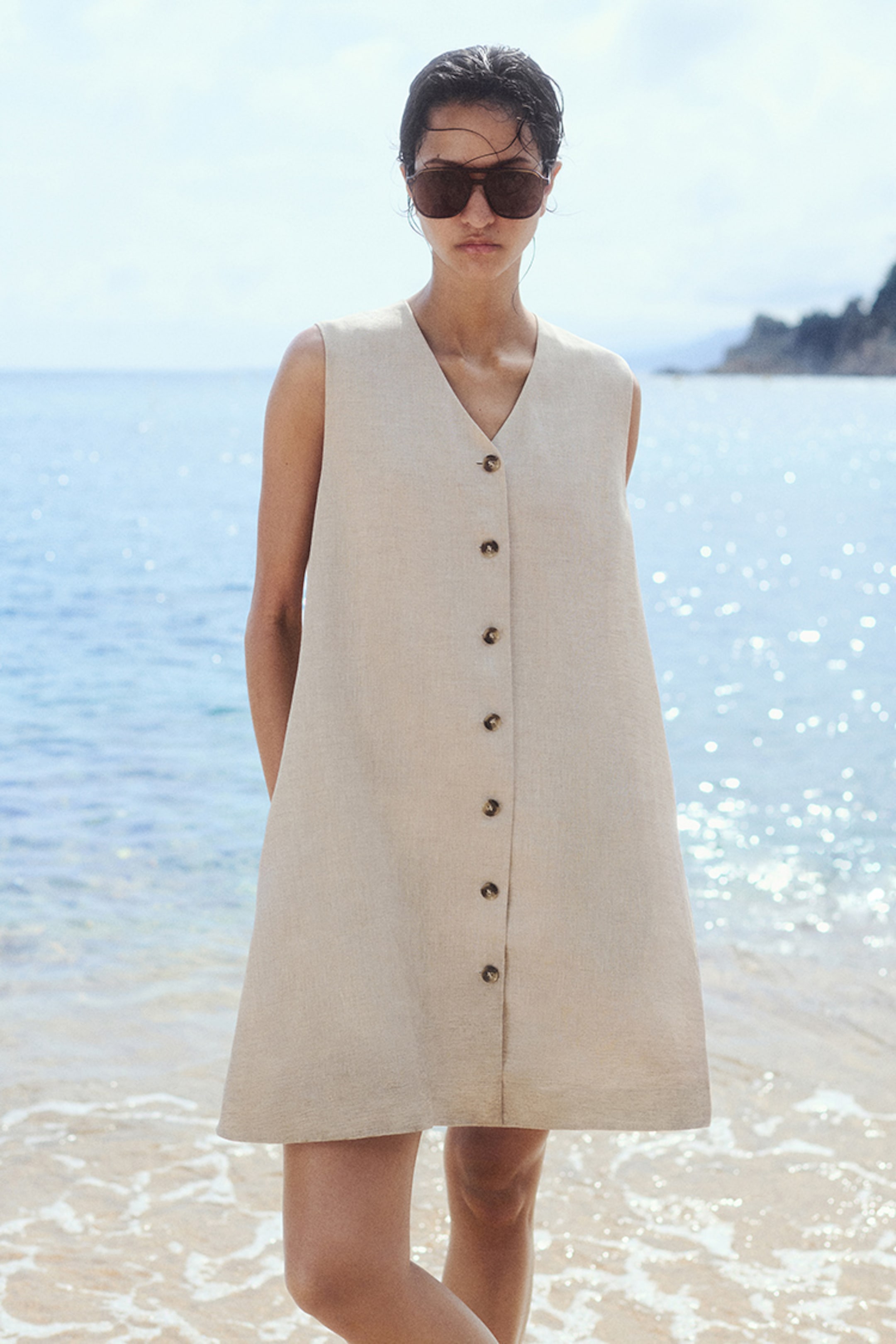 Woman in a beige sleeveless button-up dress and sunglasses stands on a sunny beach with the sea in the background.