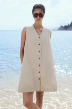 Placeholder for: Woman in a beige sleeveless button-up dress and sunglasses stands on a sunny beach with the sea in the background.