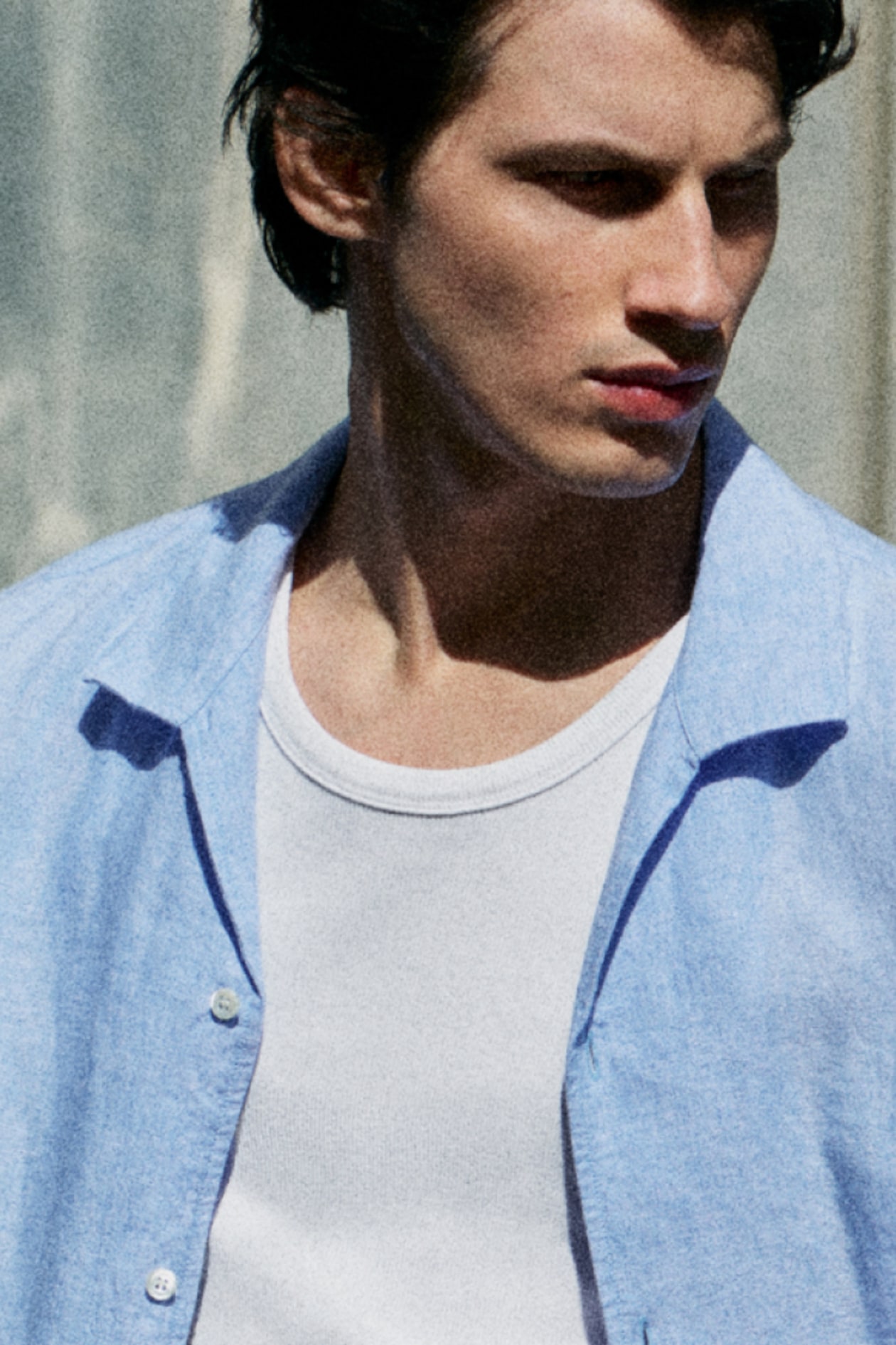 Man in a white undershirt and light blue button-up shirt, looking to the side, appears contemplative in natural lighting.