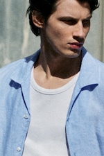 Placeholder for: Man in a white undershirt and light blue button-up shirt, looking to the side, appears contemplative in natural lighting.
