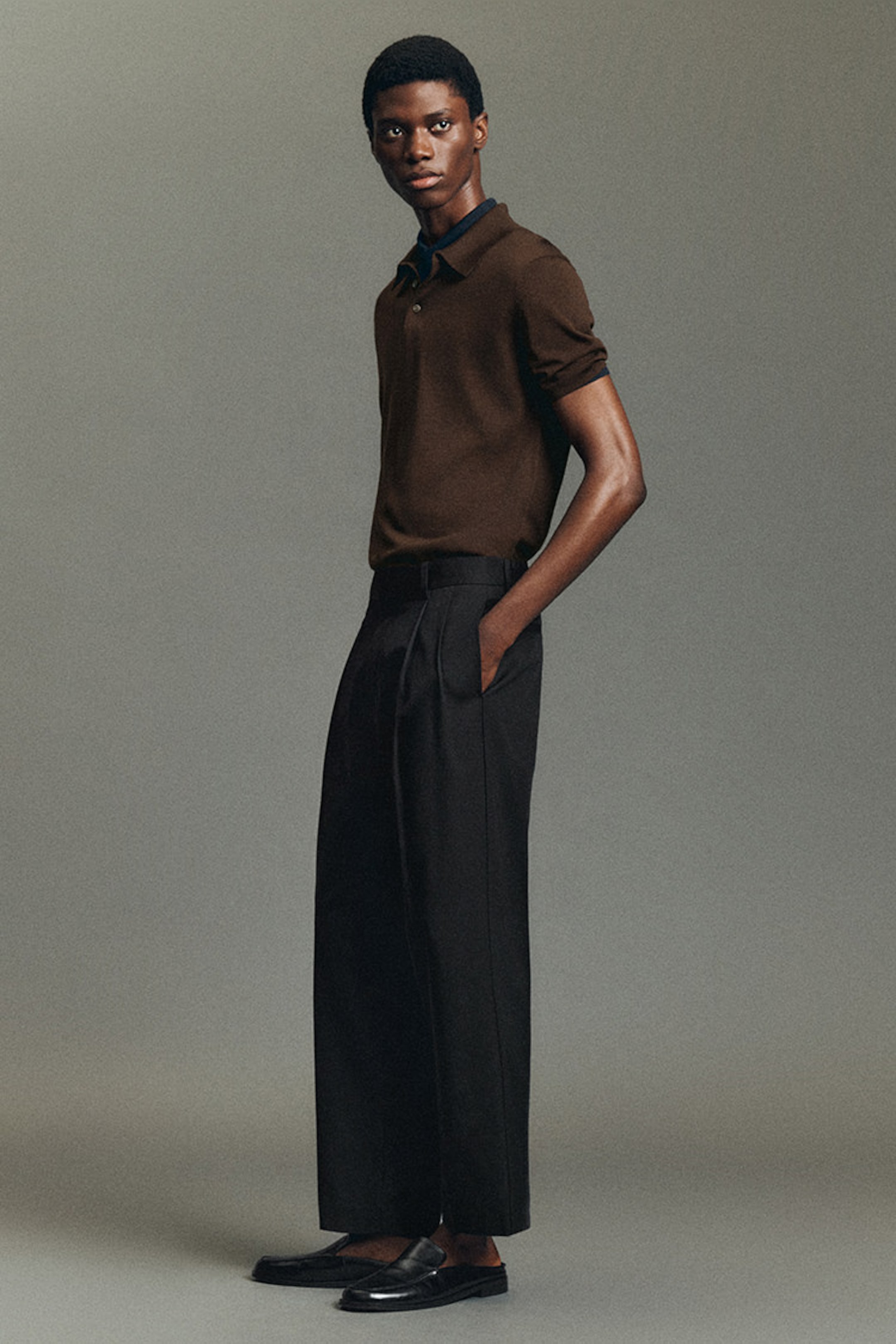 A person in a brown polo shirt and black wide-leg trousers stands against a neutral background, looking to the side with hands in pockets.