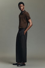 Placeholder for: A person in a brown polo shirt and black wide-leg trousers stands against a neutral background, looking to the side with hands in pockets.