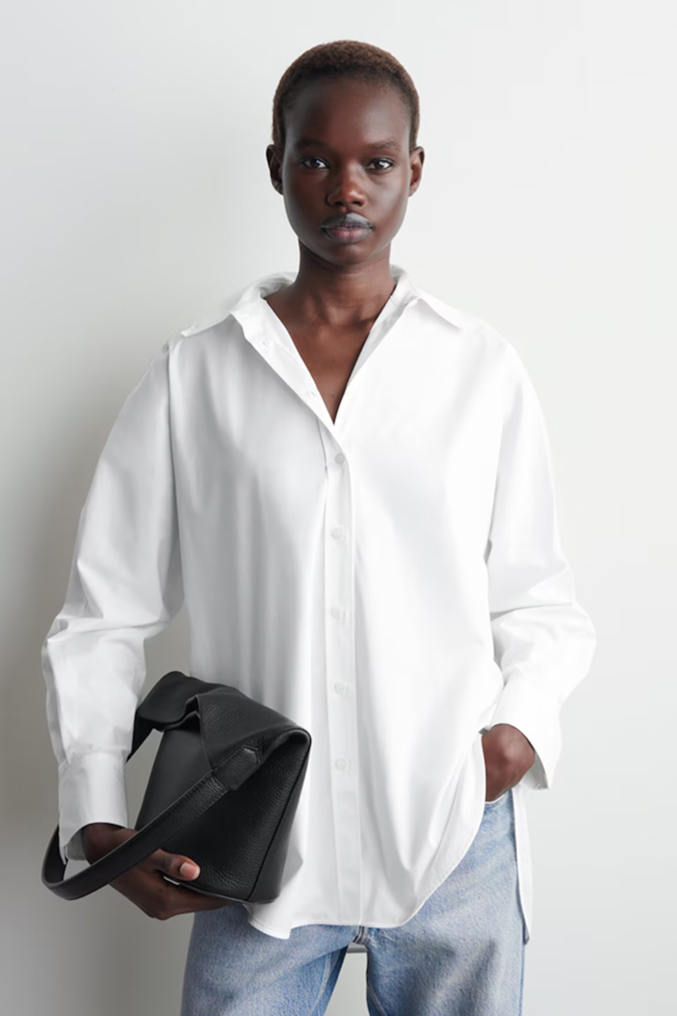 Person wearing a white button-up shirt and jeans, holding a black bag, standing against a plain light background.