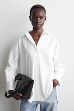 Placeholder for: Person wearing a white button-up shirt and jeans, holding a black bag, standing against a plain light background.