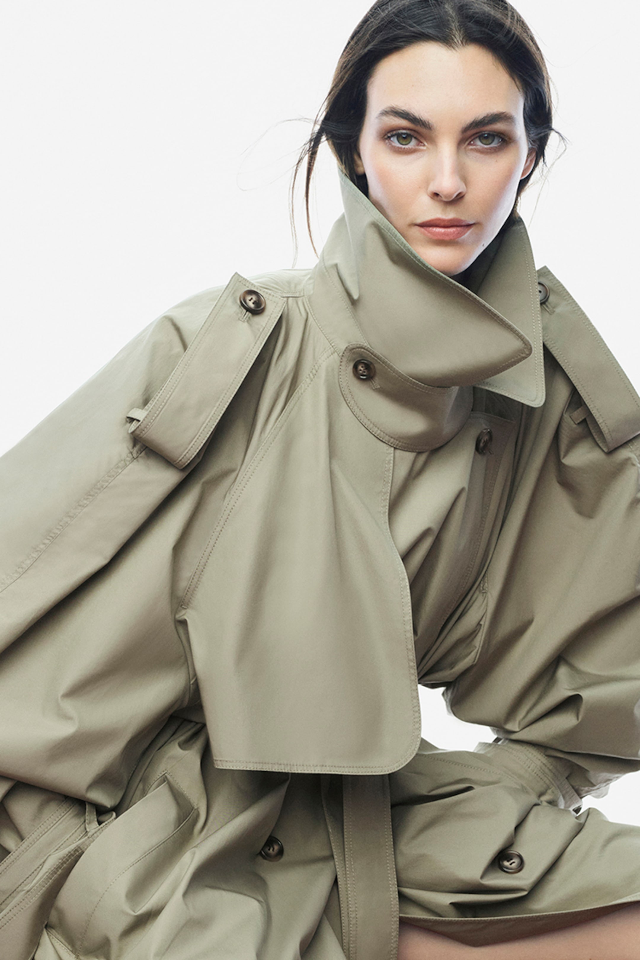 Woman in a beige oversized trench coat with high collar, looking at the camera against a plain white background.