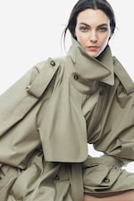 Placeholder for: Woman in a beige oversized trench coat with high collar, looking at the camera against a plain white background.