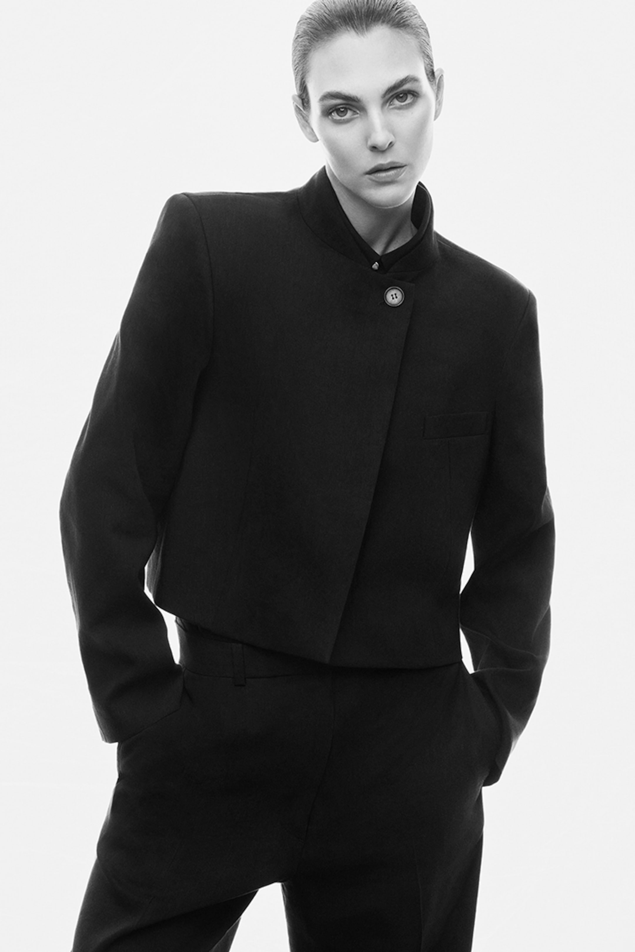 Person in a black, buttoned suit with a stand-up collar, hands in pockets, gazing confidently at the camera against a white background.