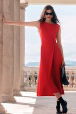 Placeholder for: Woman in a sleeveless red dress and sunglasses leans against a column, holding a black bag, with a scenic view in the background.