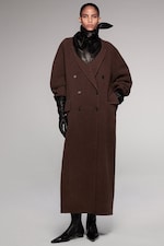 Placeholder for: A person models a long brown coat with black buttons, wearing black gloves, black scarf, and black pointed shoes, set against a plain background.