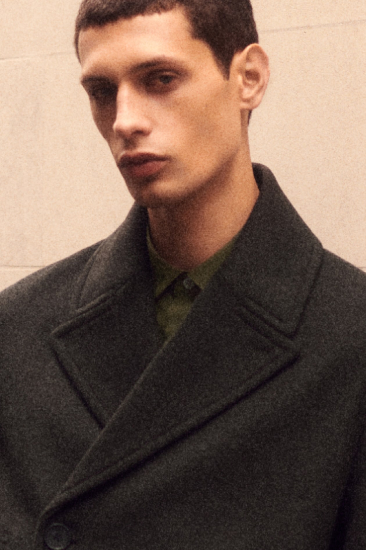Person wearing a dark, oversized coat and green shirt, standing against a plain background, looking slightly to the side.
