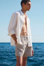 Placeholder for: Man in white shirt and striped shorts stands by the sea on a sunny day, gazing into the distance.