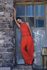 Placeholder for: Person in a sleeveless red outfit and black sandals leaning against a rustic stone and metal wall, posing outdoors casually.