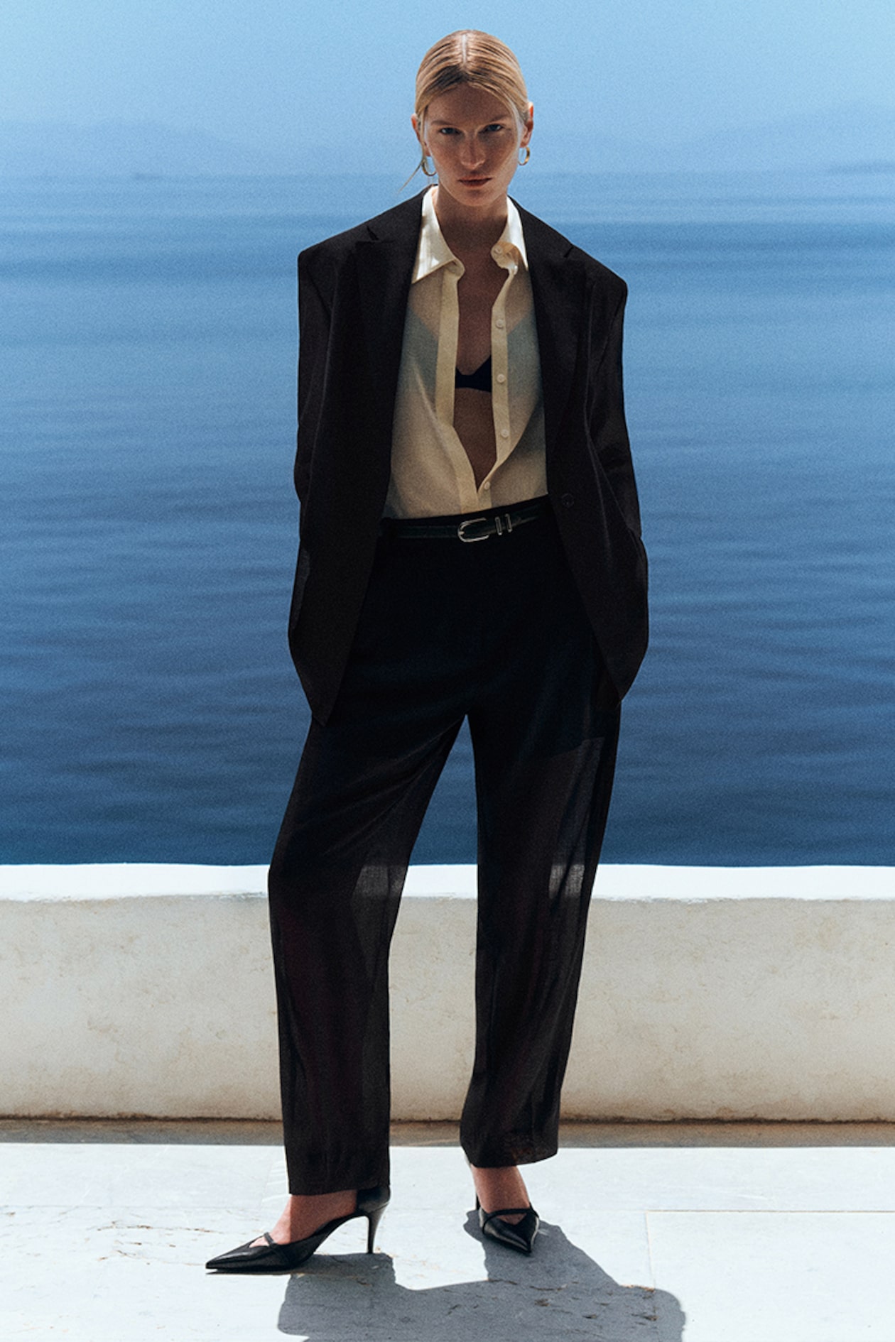 A person in a black suit, open shirt, and heels stands confidently by the sea on a sunny day, with a serene blue ocean backdrop.