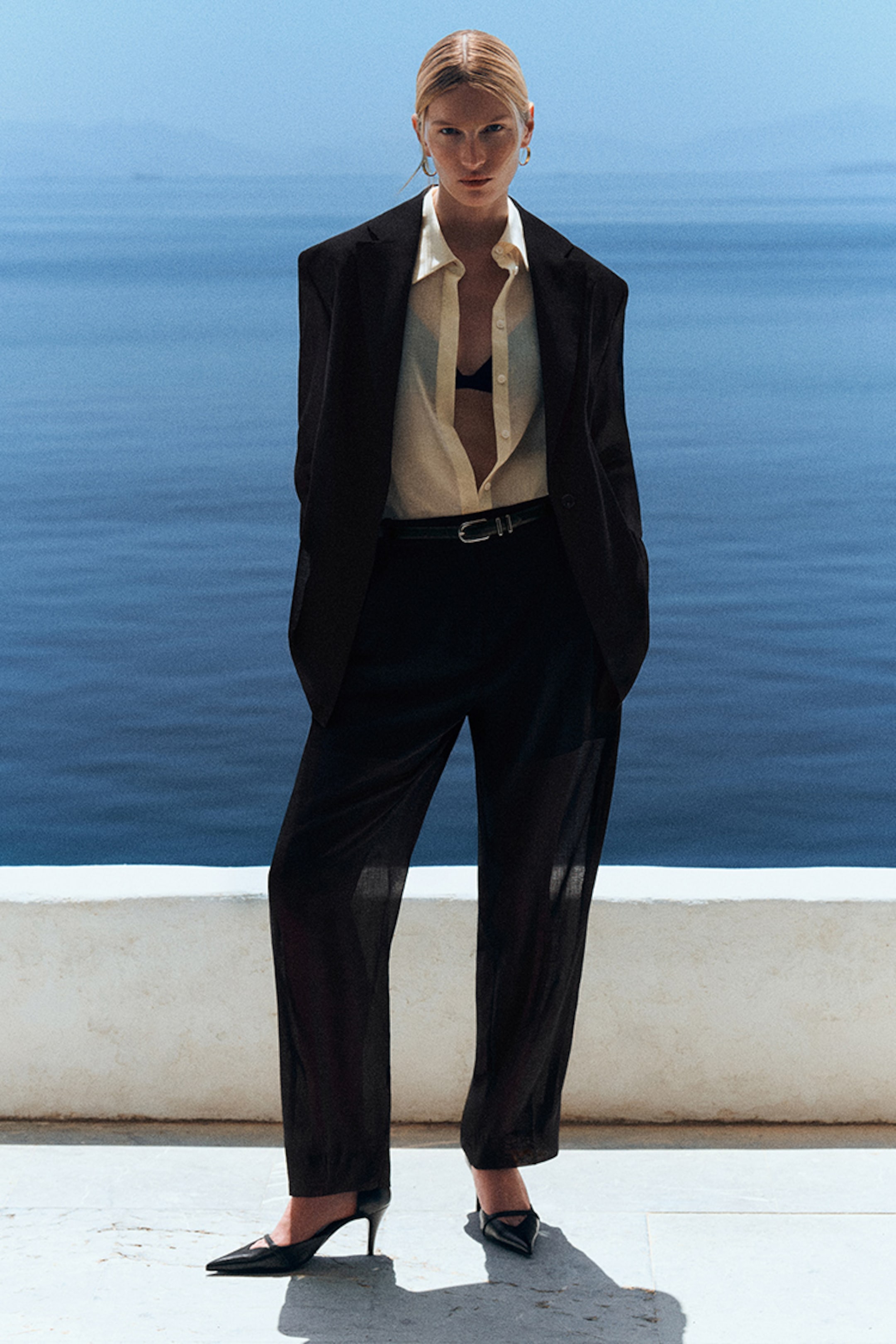 A person in a black suit, open shirt, and heels stands confidently by the sea on a sunny day, with a serene blue ocean backdrop.