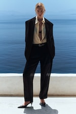Placeholder for: A person in a black suit, open shirt, and heels stands confidently by the sea on a sunny day, with a serene blue ocean backdrop.