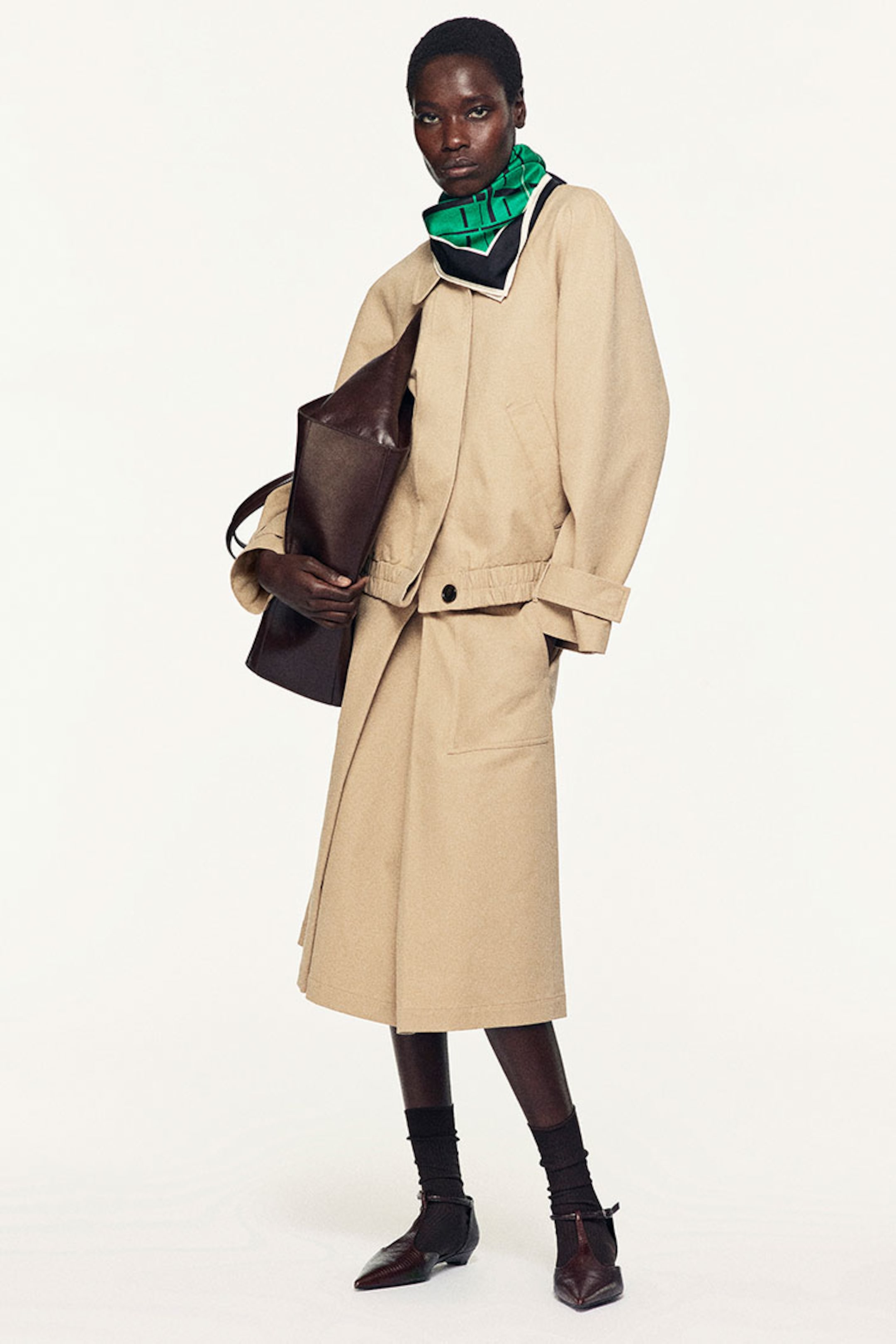 Person in a beige outfit with a green scarf, holding a brown bag, and wearing brown shoes against a plain background.