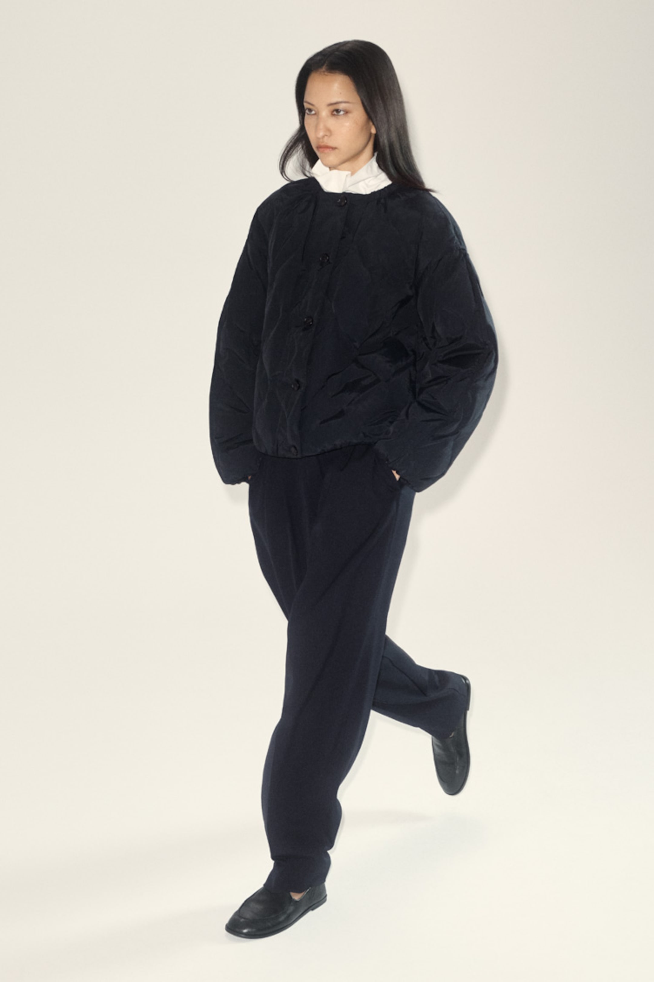 A person in a black quilted jacket, white shirt, and black pants walks confidently against a light background.