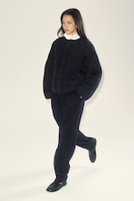 Placeholder for: A person in a black quilted jacket, white shirt, and black pants walks confidently against a light background.