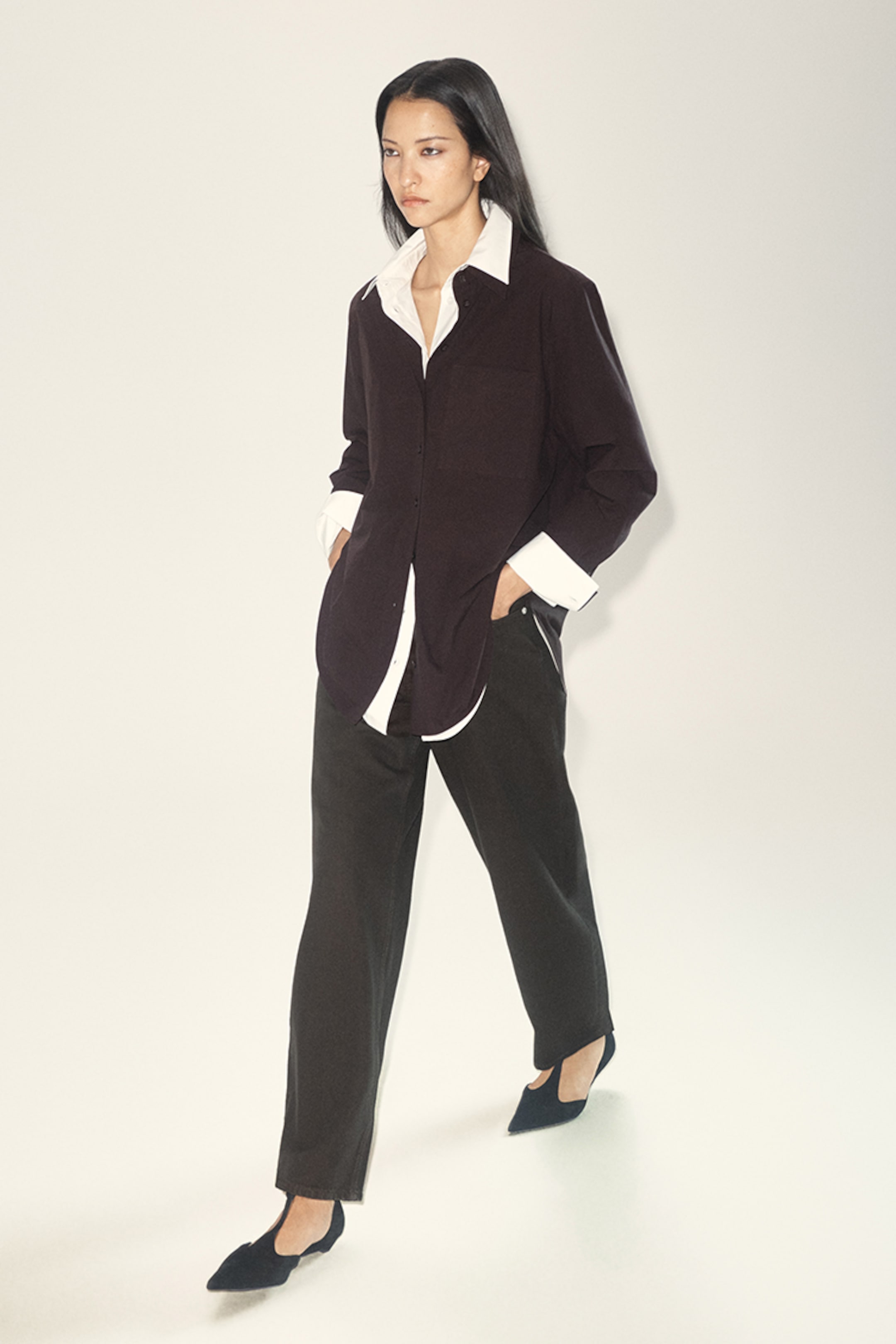 Woman walking in a minimalist setting, wearing a black blazer over a white shirt, black pants, and black pointed shoes.