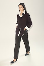 Placeholder for: Woman walking in a minimalist setting, wearing a black blazer over a white shirt, black pants, and black pointed shoes.