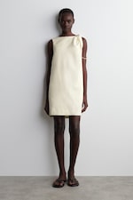 Placeholder for: A person stands against a white wall wearing a sleeveless, light yellow dress with a shoulder tie, paired with black sandals.