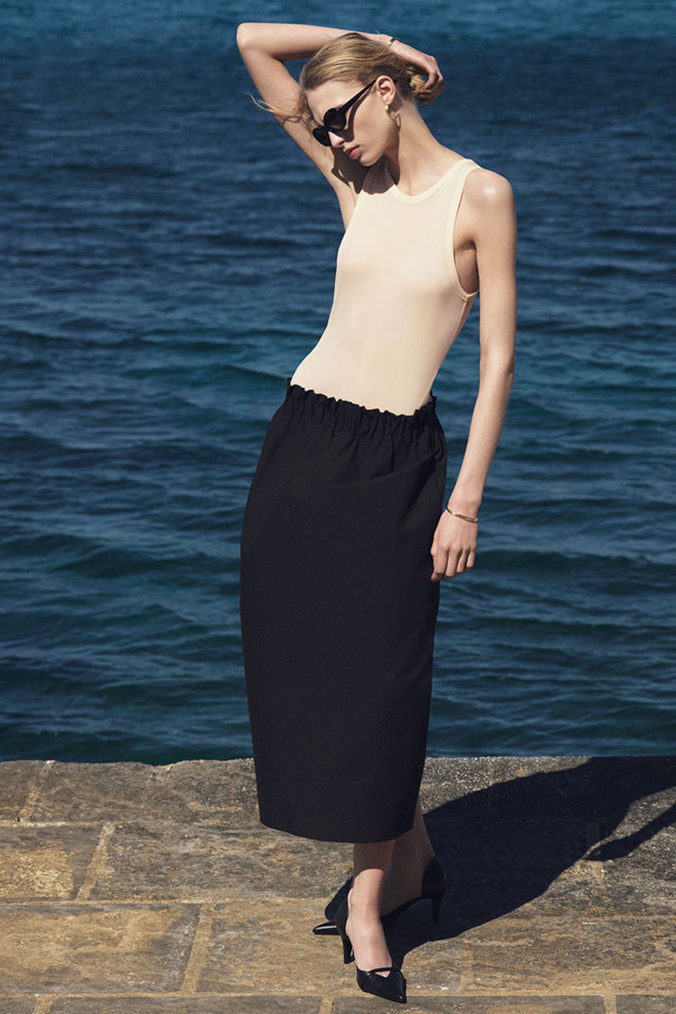 Woman in a sleeveless top and black skirt poses on a stone pathway by the sea, wearing sunglasses and black heels.