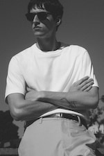 Placeholder for: Black and white image of a person wearing sunglasses and a white t-shirt, arms crossed, with a tattoo on the forearm.