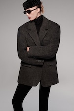 Placeholder for: Person wearing a large dark gray coat, black turtleneck, sunglasses, and a textured hat, with arms crossed in a fashion pose.