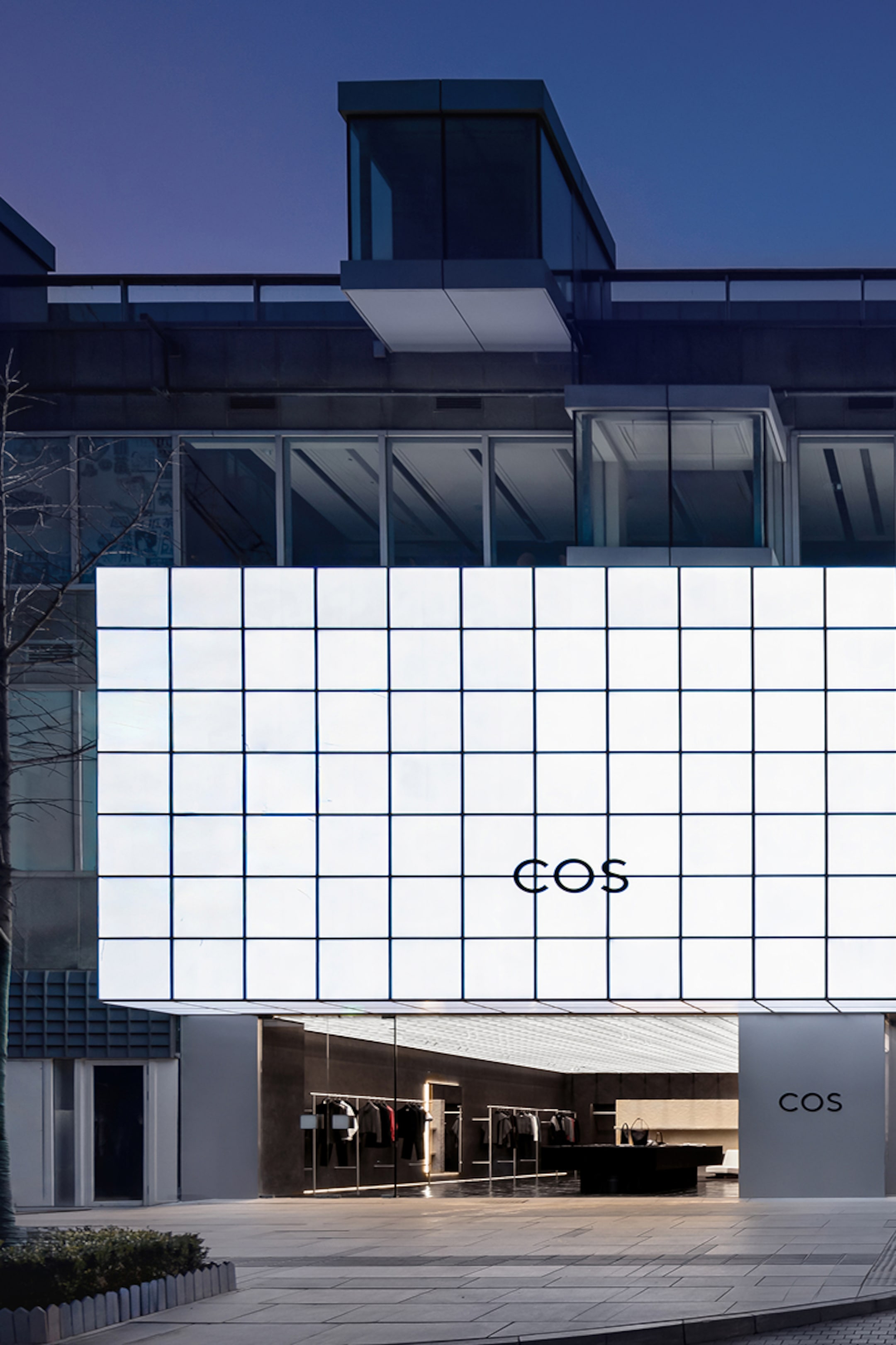Modern building with a large, illuminated grid facade and "COS" logo. Store entrance below with visible interior displaying clothing.